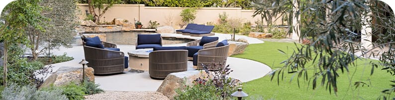 Wicker chairs positions in a circle around a fire pit, in a backyard with green grass, stone boulders, and a pool.