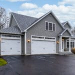 Multi-story home with grey shingles, grey front, and white garage doors. Home located near tall wooded area.