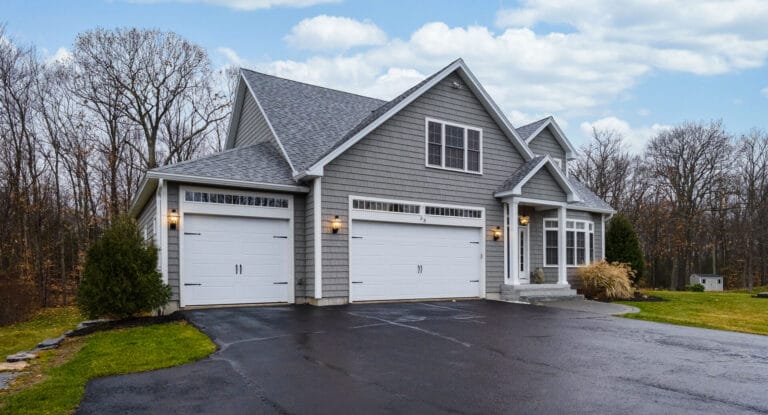 Multi-story home with grey shingles, grey front, and white garage doors. Home located near tall wooded area.