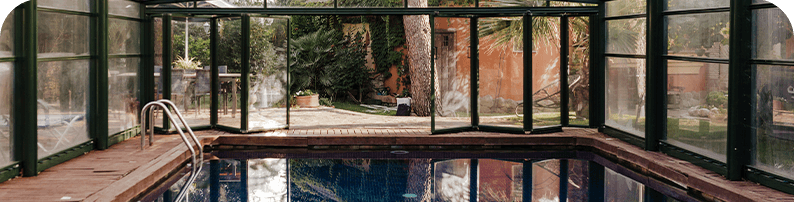 An indoor pool with glass walls and doors, seamlessly connecting to a lush outdoor garden with greenery and terracotta-colored walls
